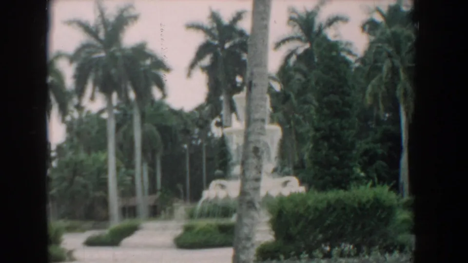 1958 South Carolina home-movie frame showing a white fountain with water sprays, hedges, palm trees, and soft color 8mm texture.