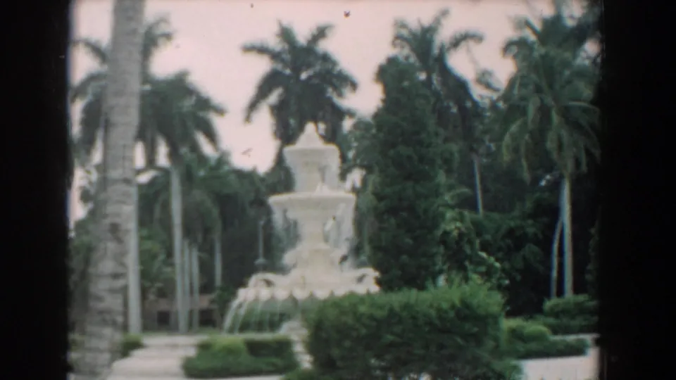 1958 South Carolina frame of a white fountain and palm-lined garden, with hedges in front and a dark vertical film edge at left.