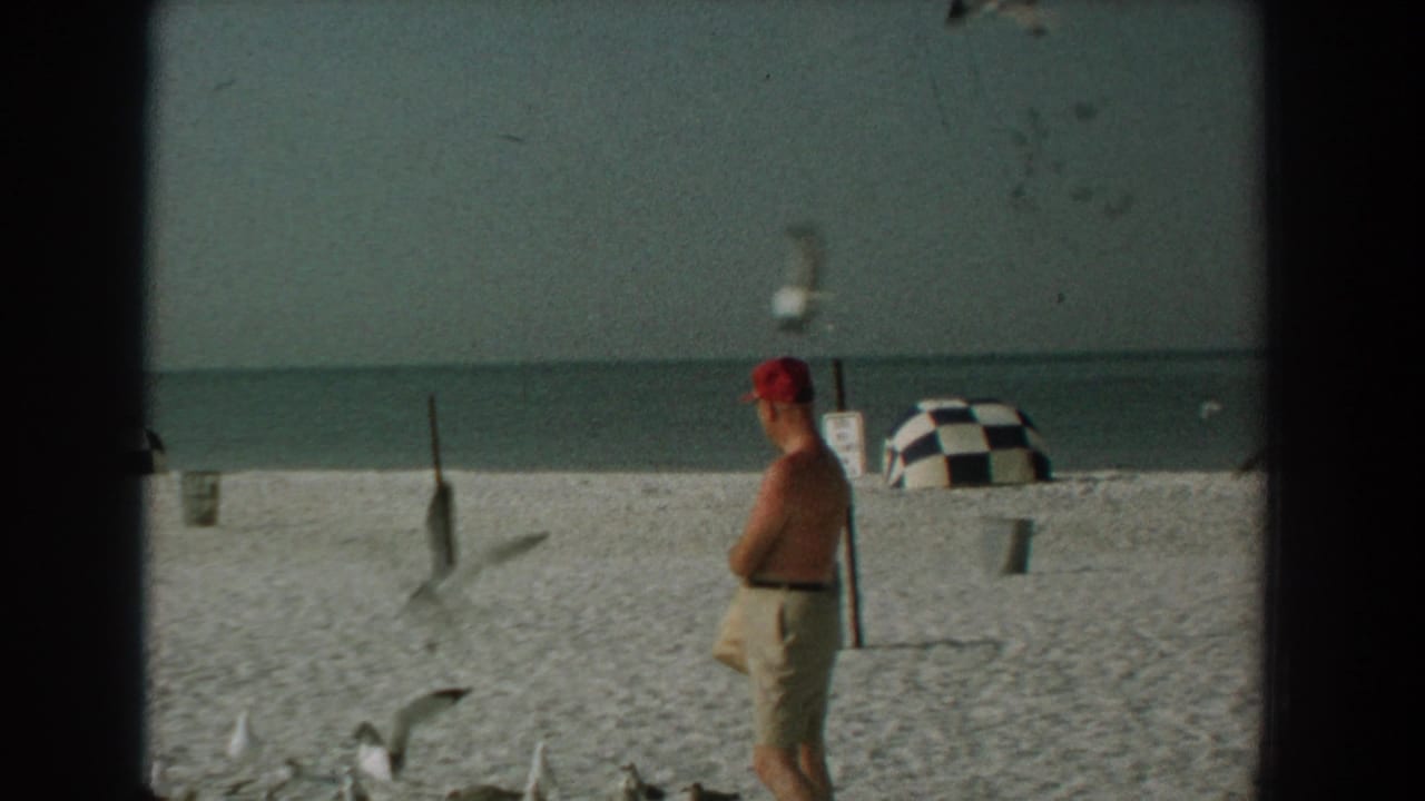 In supported 1958 Yorktown beach footage, a shirtless man in a red cap stands near gulls with the ocean and a checkered cabana behind him.
