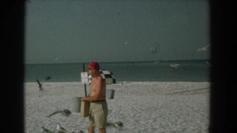 A shirtless man in a red cap appears to hold a paper bag while gulls fly low over the 1958 Yorktown beach scene.