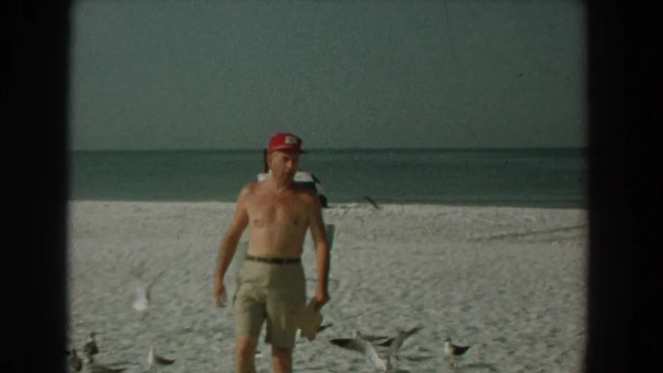 A shirtless man in a red cap and khaki shorts stands among gulls on the 1958 Yorktown beach.