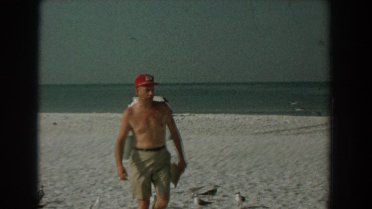 Shirtless man in red cap walking on a sandy beach with seagulls and ocean behind him.