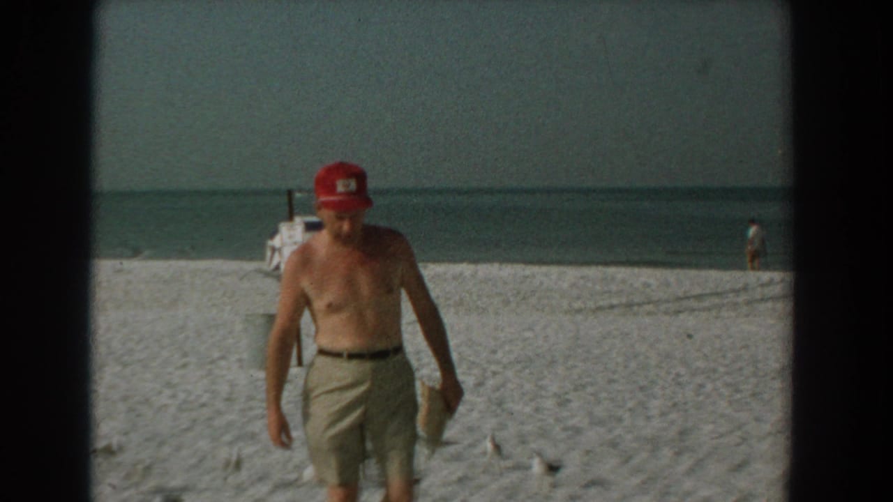 Man in red cap walking closer to camera on beach with seagulls and distant shoreline.