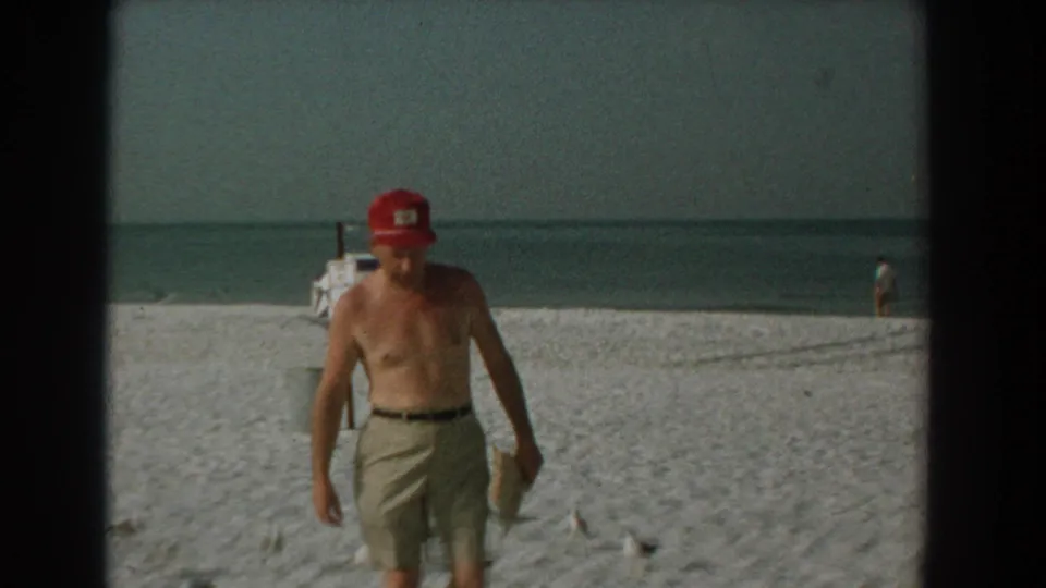A shirtless man in khaki shorts and a red cap walks along the Yorktown beach as gulls stand on the sand nearby.