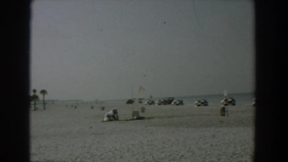 Wide 1958 Yorktown beach view with umbrellas or shade structures, parked cars, open sand, and the ocean horizon in the distance.