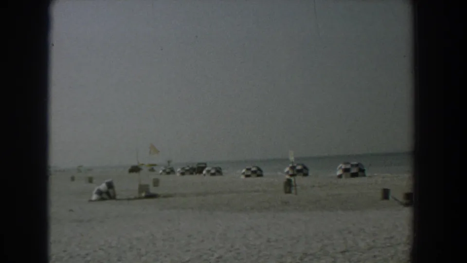 Wide Yorktown beach frame showing parked cars near the shoreline, simple umbrellas, open sand, and a distant standing figure.