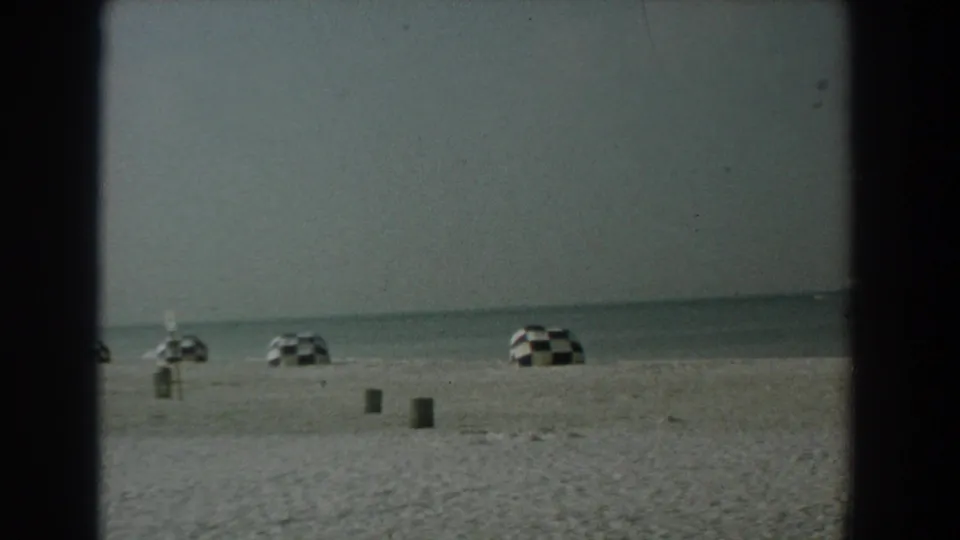 1958 Yorktown shoreline with widely spaced checkered umbrellas, beach bins, and a soft ocean horizon.