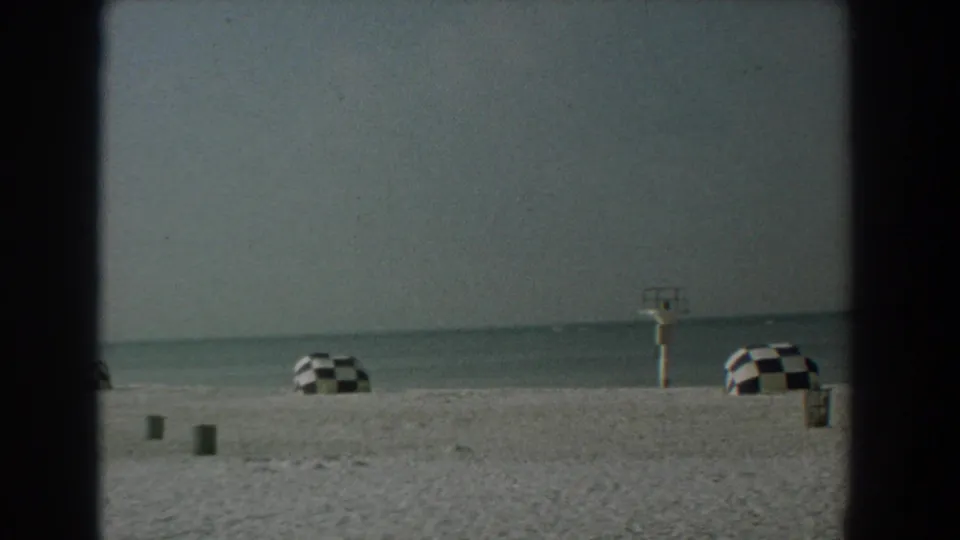 1958 Yorktown, Virginia beach view with pale sand, calm water, checkered umbrellas, and a small lifeguard stand under a gray sky.