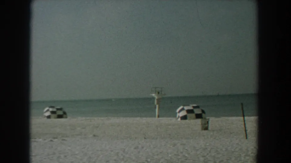 1958 Yorktown, Virginia shoreline with checkered umbrellas spaced across pale sand and a small lifeguard stand near calm water.