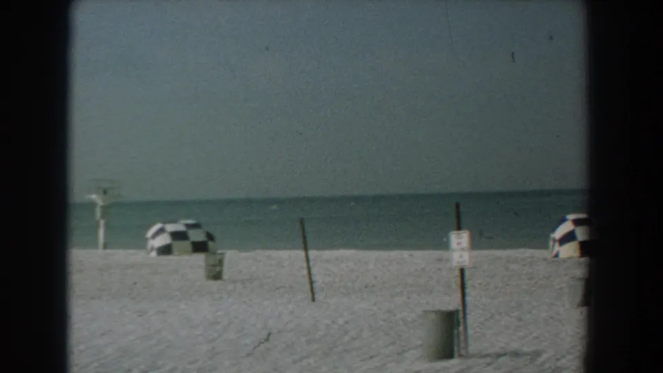 1958 Yorktown, Virginia beach view with checkered umbrellas along the sand, a leaning post, a small sign, and calm water under gray sky.