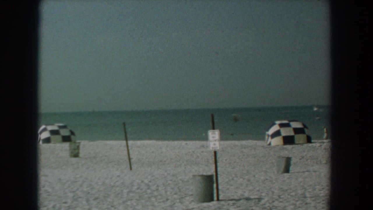 Wide beach with calm ocean, two checkered umbrellas, a signpost, and a trash can.