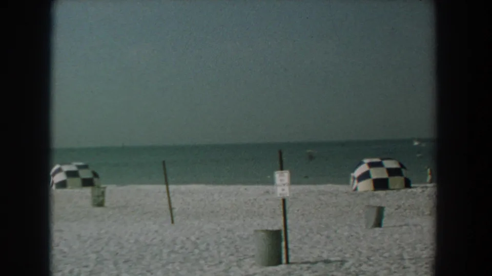 1958 Yorktown beach view with pale sand, calm water, two black-and-white checkered umbrellas, a signpost, and a trash can.