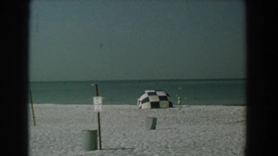 Yorktown, Virginia beach scene with a checkered umbrella near the shoreline, a signpost, a trash can, and small seagulls over calm water.
