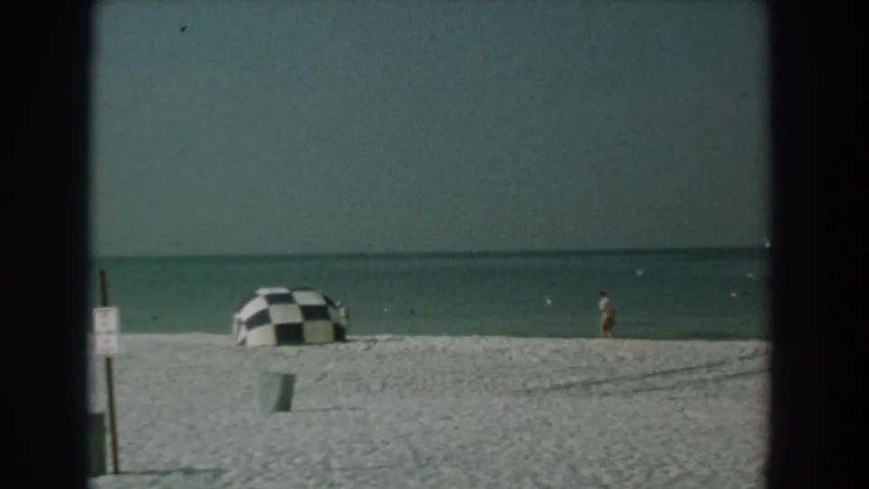 1958 Yorktown beach frame showing a checkered umbrella, signpost, trash can, calm ocean, and a distant person walking near the water.