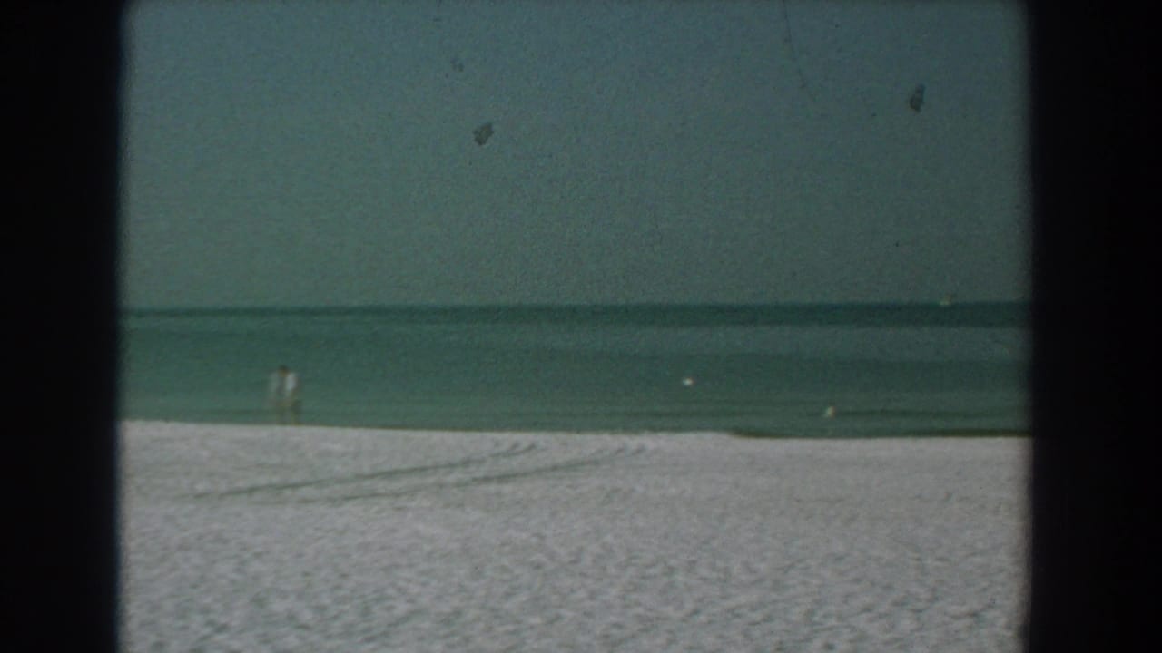 White sand beach with calm water, pale sky, distant birds, and dark 8mm frame borders.