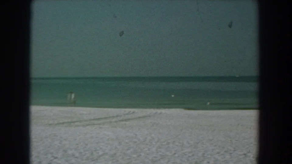 1958 Yorktown, Virginia beach view with pale sand, blue-green water, a faint distant figure, birds, and dark 8mm film borders.