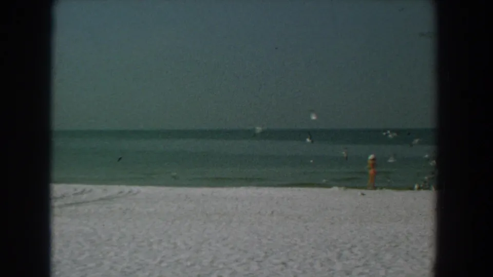 1958 Yorktown beach frame with a standing beachgoer near the water, birds overhead, pale sand, and a calm horizon.