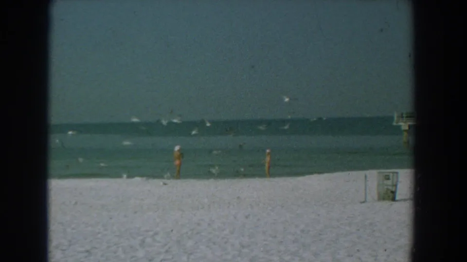 1958 Yorktown beach view with scattered people, birds near the water, a shoreline bin at right, and visible 8mm frame edges.