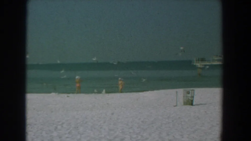 1958 Yorktown, Virginia beach footage shows two women in swimwear near calm water with gulls above and a distant pier at right.