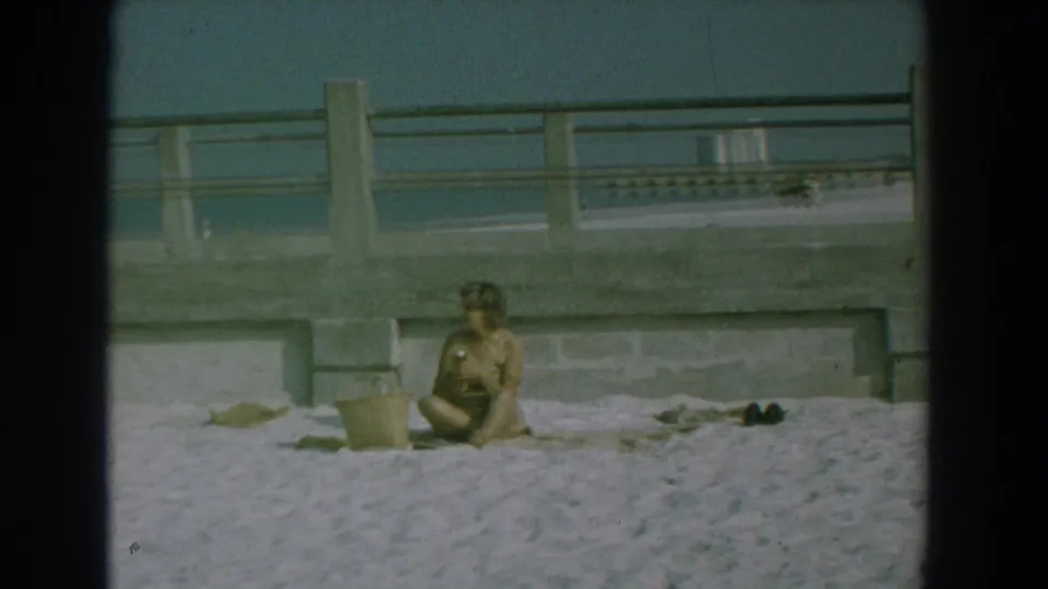 A woman sits on the Yorktown beach sand beneath a concrete seawall, with a bucket, shoes, and small beach items arranged nearby.