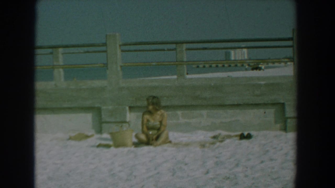 Woman seated on a towel on a sandy beach near a bucket and concrete railing.
