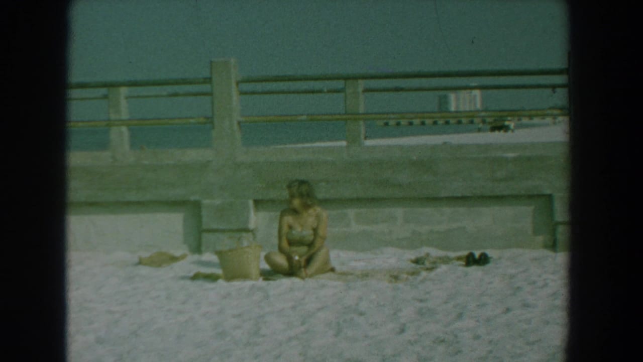 The woman remains seated on the Yorktown beach, looking slightly aside near a towel, bucket, concrete railing, and water in the background.