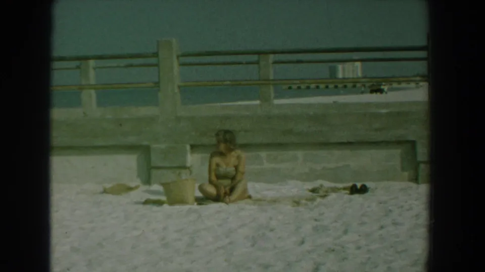 A 1958 home-movie frame shows the woman seated low on the sand near a bucket, with a concrete waterfront barrier and water behind her.