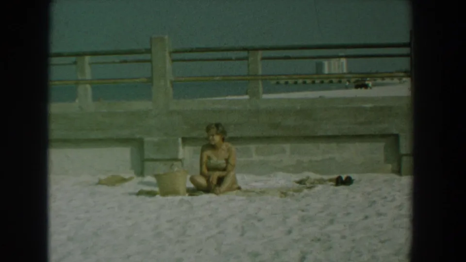 The woman sits on the sandy Yorktown waterfront beside a towel and bucket, with dark shoes or belongings nearby and railing beyond.