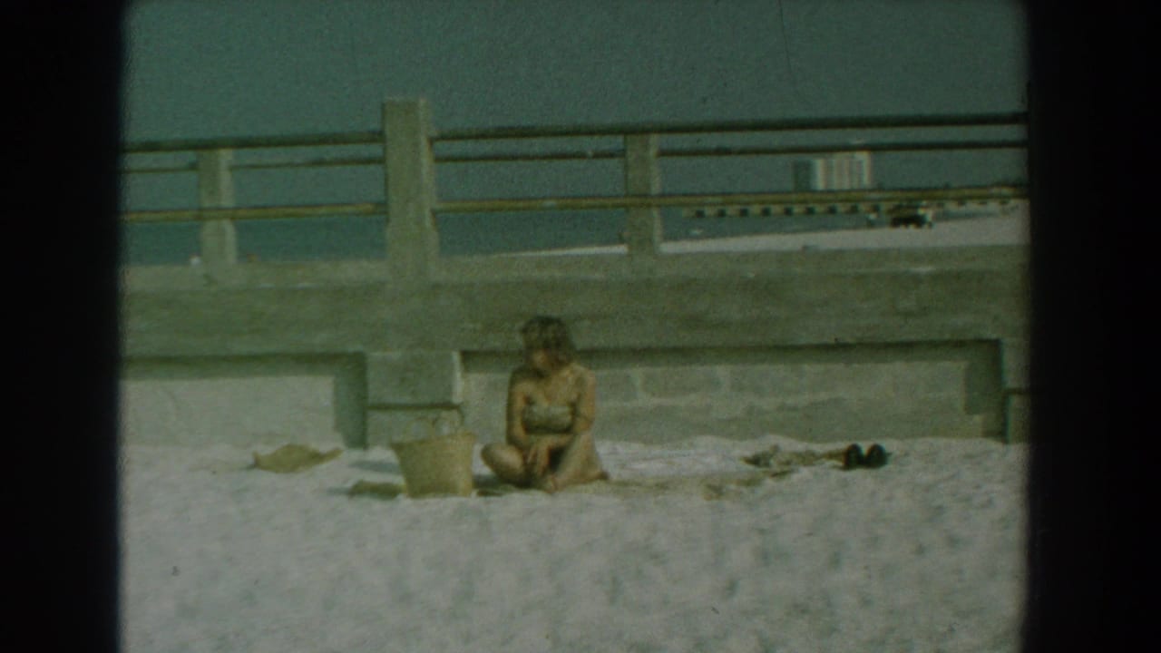 Woman seated on sand near a bucket, towel, shoes, concrete railing, and ocean.