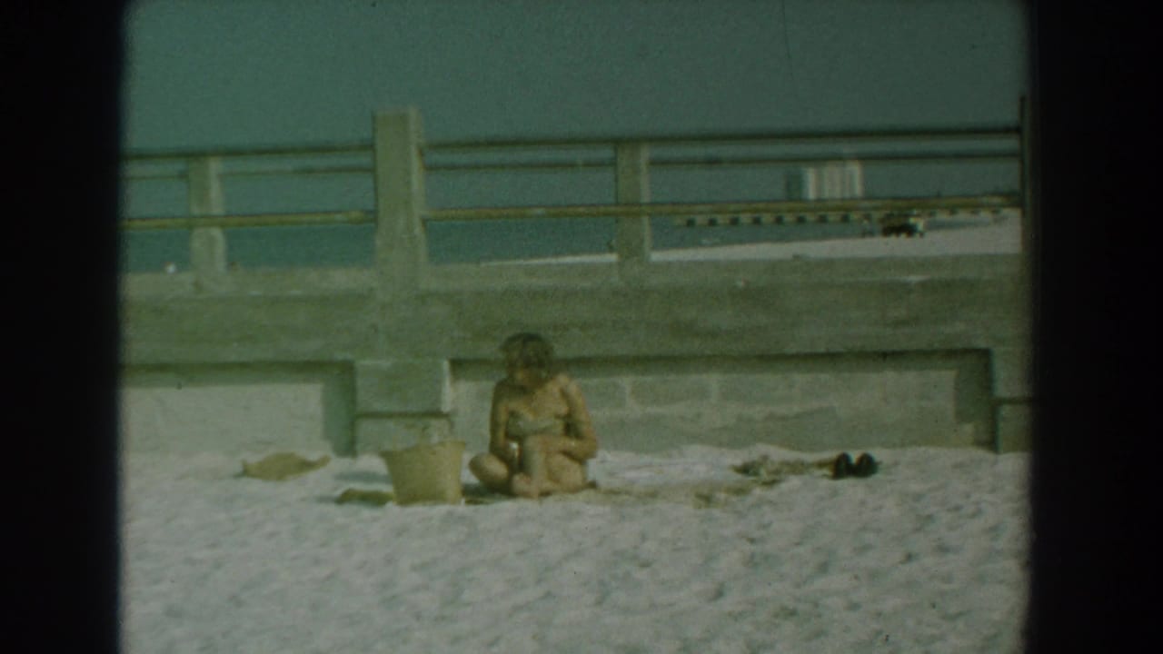 Woman sitting on sand beside beach items with a concrete railing and water behind.