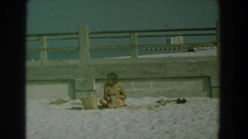 A woman remains seated on the sand in the Yorktown waterfront view, with a bucket to one side and shoes or dark items nearby.