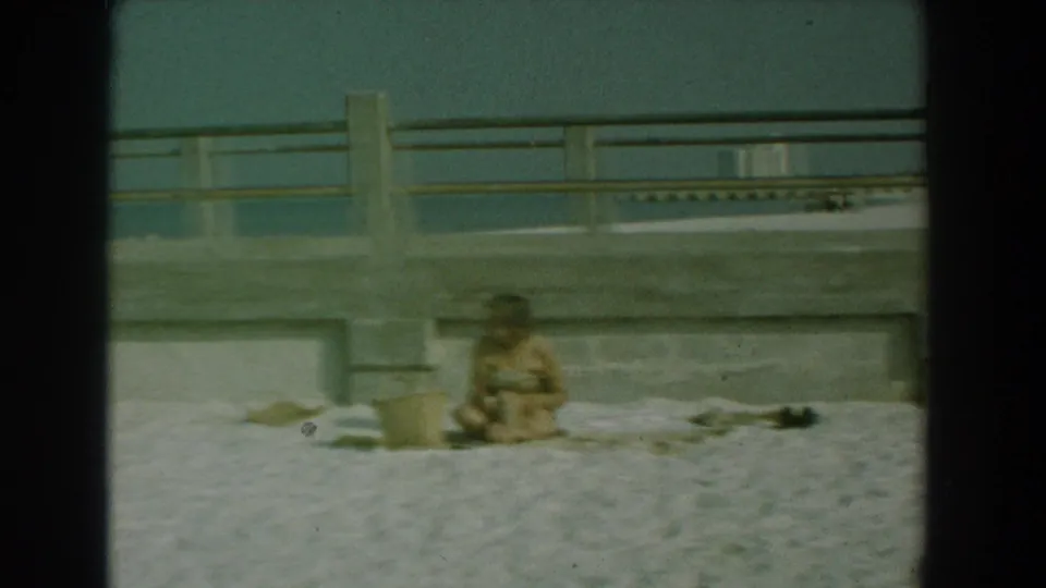 1958 Yorktown, Virginia beach scene with a woman seated on pale sand near a concrete pier wall under hazy daylight.