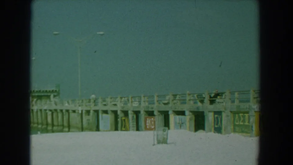 1958 Yorktown, Virginia frame showing painted Greek letter signs along the pier wall, with beach sand and light poles visible.