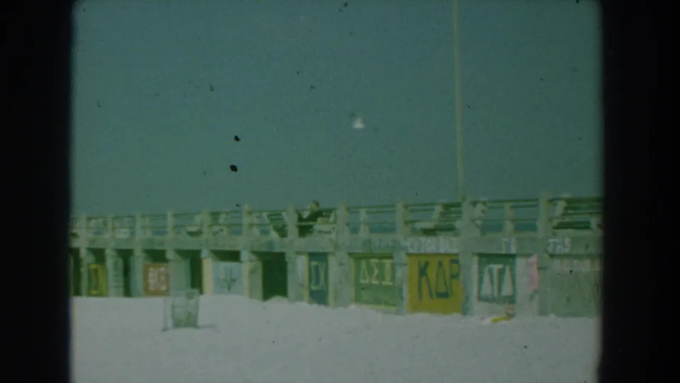 1958 Yorktown, Virginia 8mm frame showing a concrete beach boardwalk with painted Greek-letter signs beneath the railing.