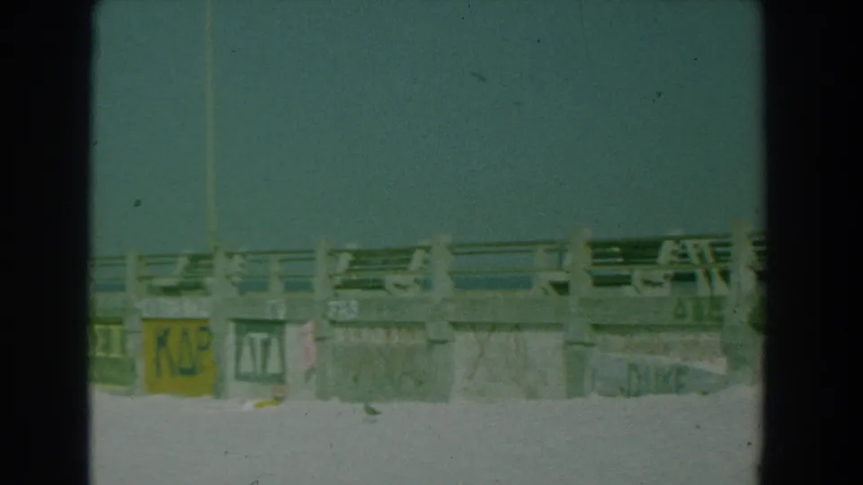 A soft 8mm view of the Yorktown beach boardwalk shows concrete railings, sand, and partially visible painted Greek-letter panels.