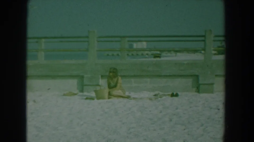 1958 Yorktown beach frame with a seated person on the sand below a concrete railing and the ocean horizon behind it.
