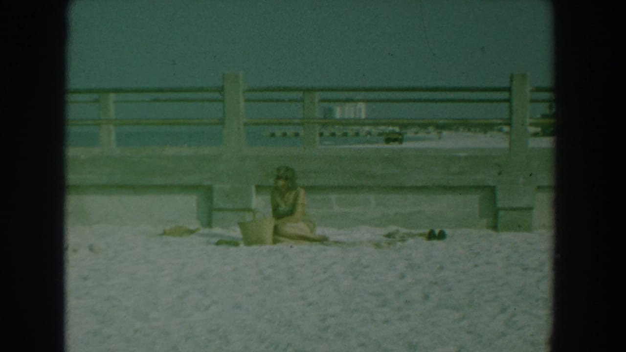 Woman seated on a sandy beach below a concrete railing with a basket beside her.