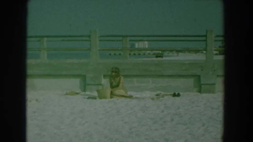 1958 Yorktown, Virginia home-movie frame showing a woman in a swimsuit sitting on sand below a concrete seaside walkway.