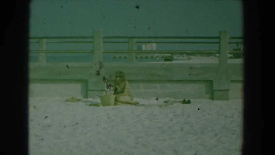 1958 Yorktown beach footage frame with a woman leaning toward a basket on sand beneath a concrete walkway.