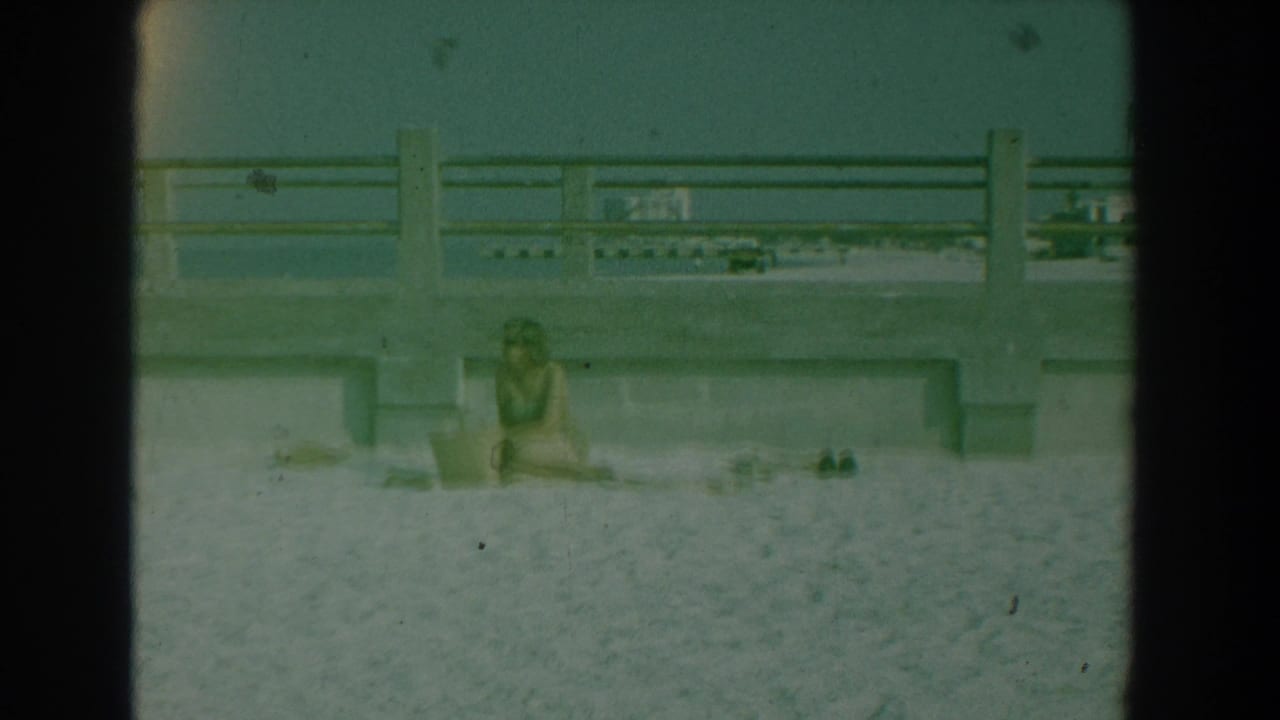 A woman sits alone on the sand at Yorktown, Virginia, framed by concrete railings in green-tinted 8mm footage.