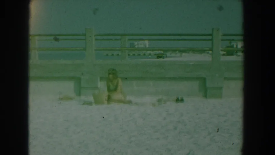 A woman sits alone on the sand at Yorktown, Virginia, framed by concrete railings in green-tinted 8mm footage.
