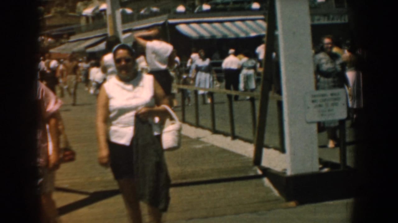 A woman in sleeveless top and skirt walks toward the camera on a 1958 Yorktown boardwalk, with other visitors behind her.