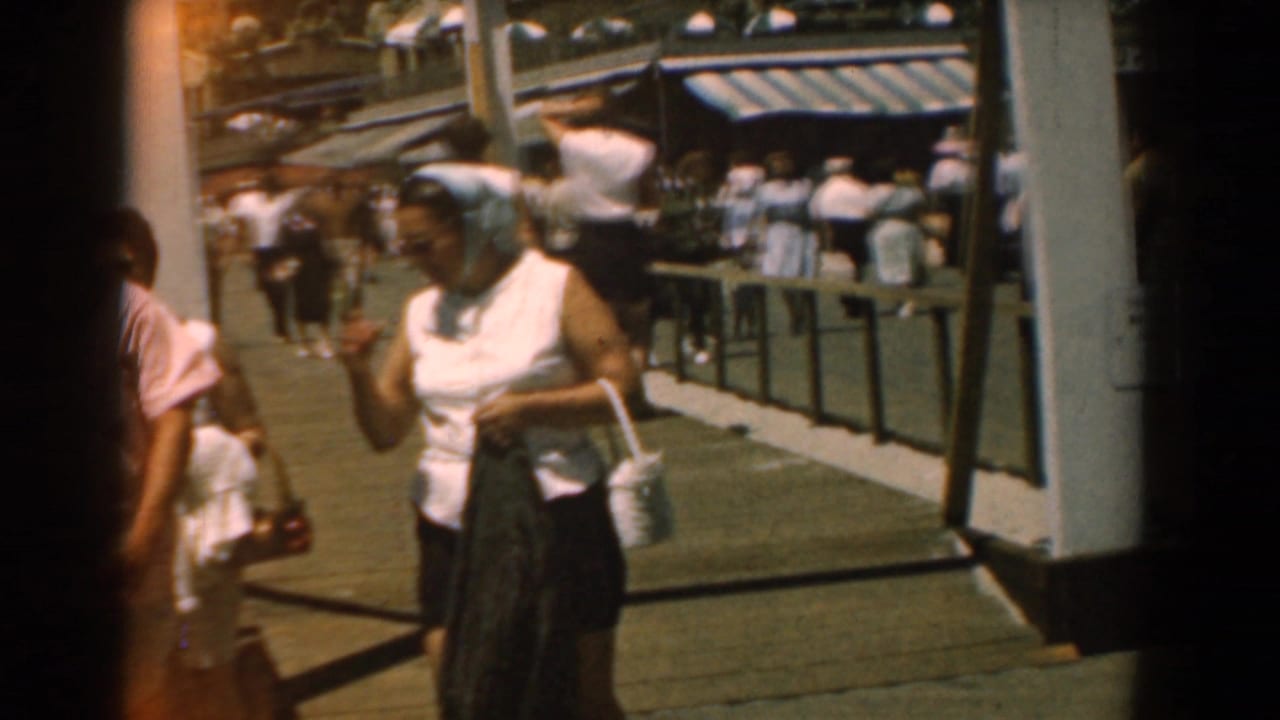 Woman in a headscarf walks toward the camera on a sunny boardwalk with pedestrians behind her.