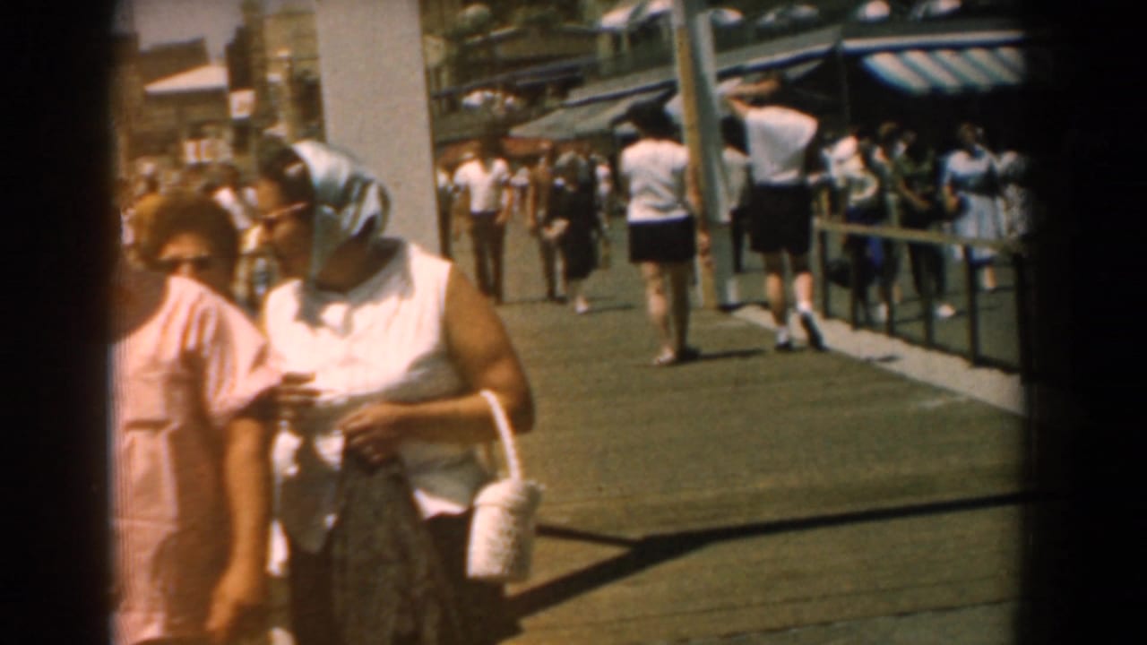 1958 Yorktown boardwalk frame showing two women close to the lens, including one in a headscarf, with pedestrians and awnings behind.