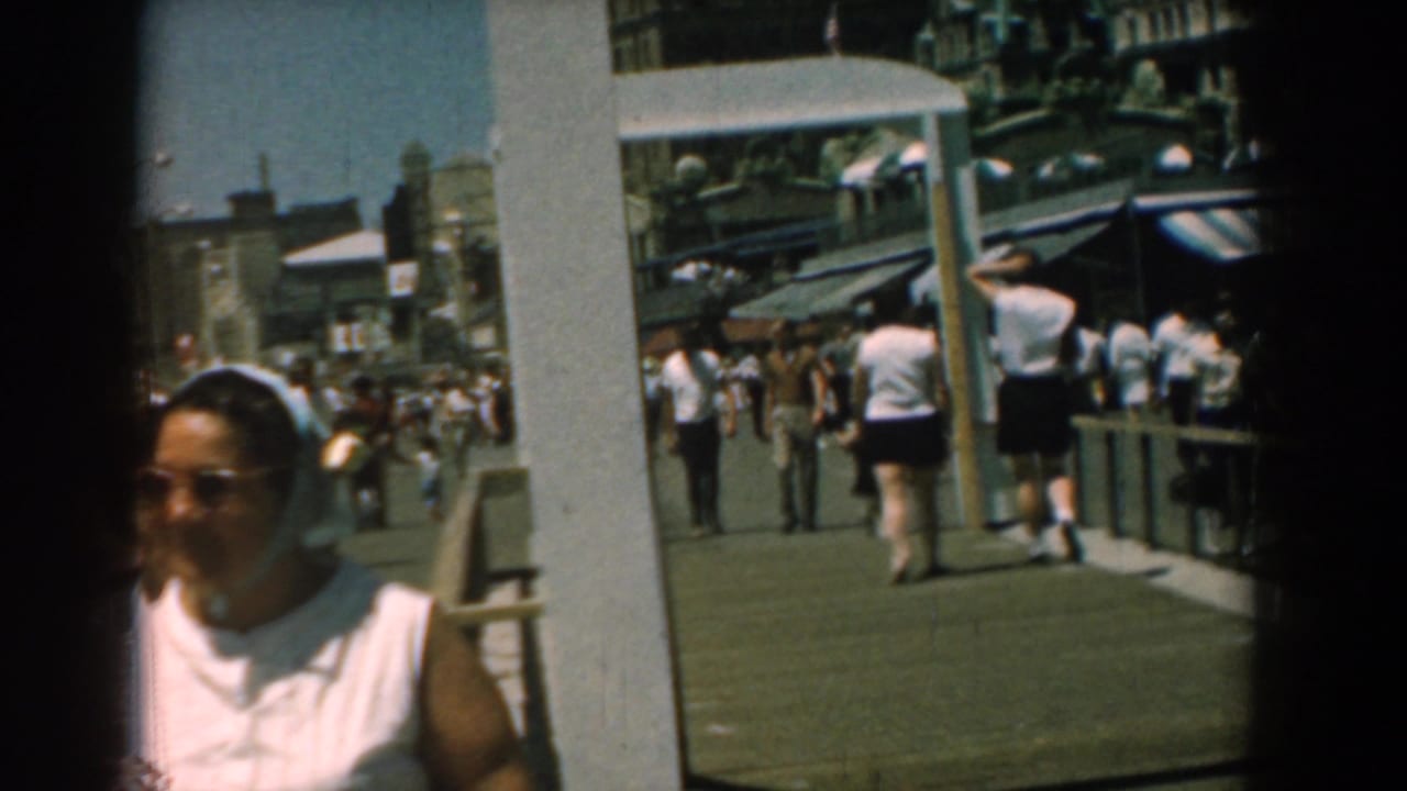Sunlit 1958 Yorktown boardwalk view with a woman partly leaving frame, visitors walking away, posts, storefronts, and striped awnings.