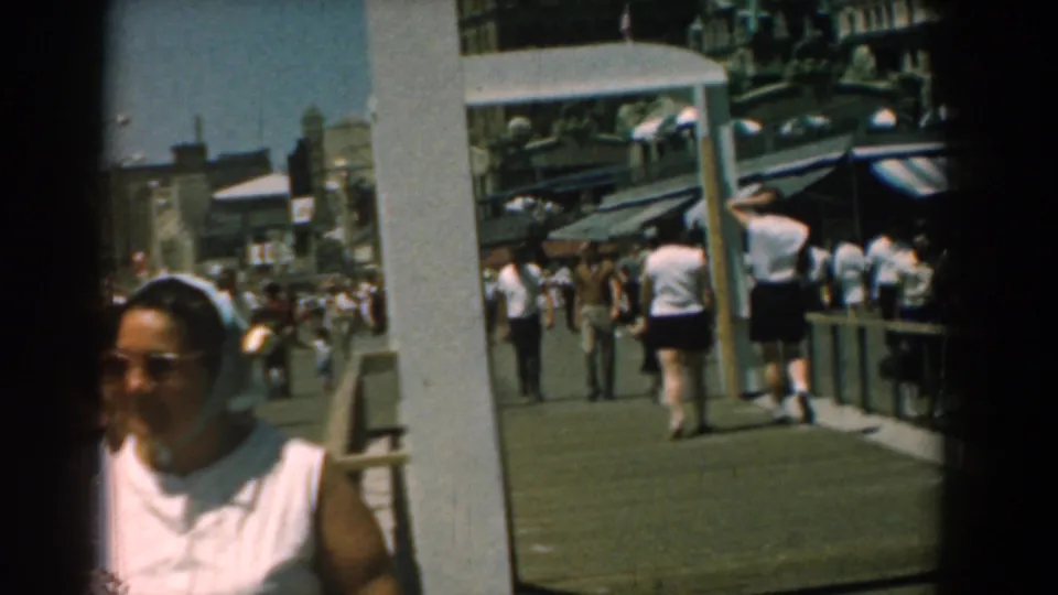 Sunlit 1958 Yorktown boardwalk view with a woman partly leaving frame, visitors walking away, posts, storefronts, and striped awnings.