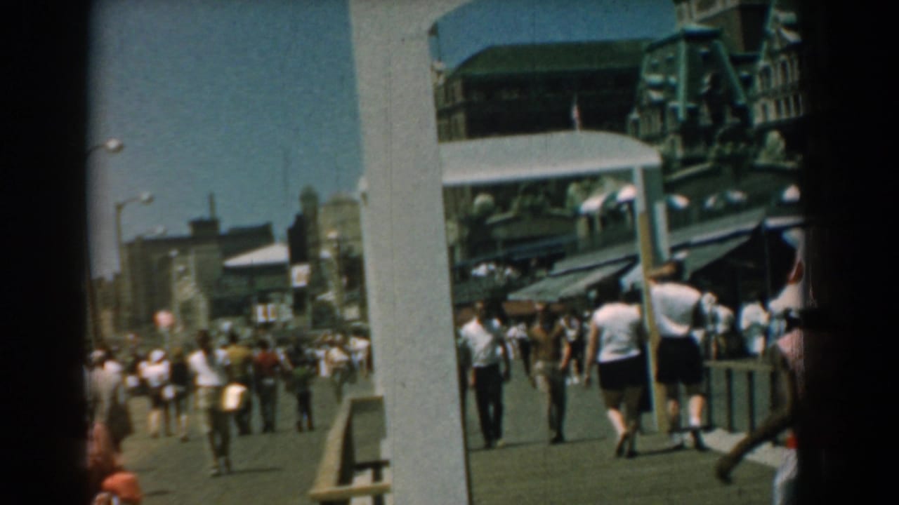 1958 Yorktown boardwalk scene with pedestrians walking away under strong sun, framed by posts, striped awnings, and waterfront storefronts.