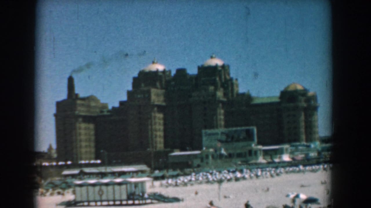 Wide beach view with umbrellas, bathers, huts, large domed buildings, and a smoking stack.