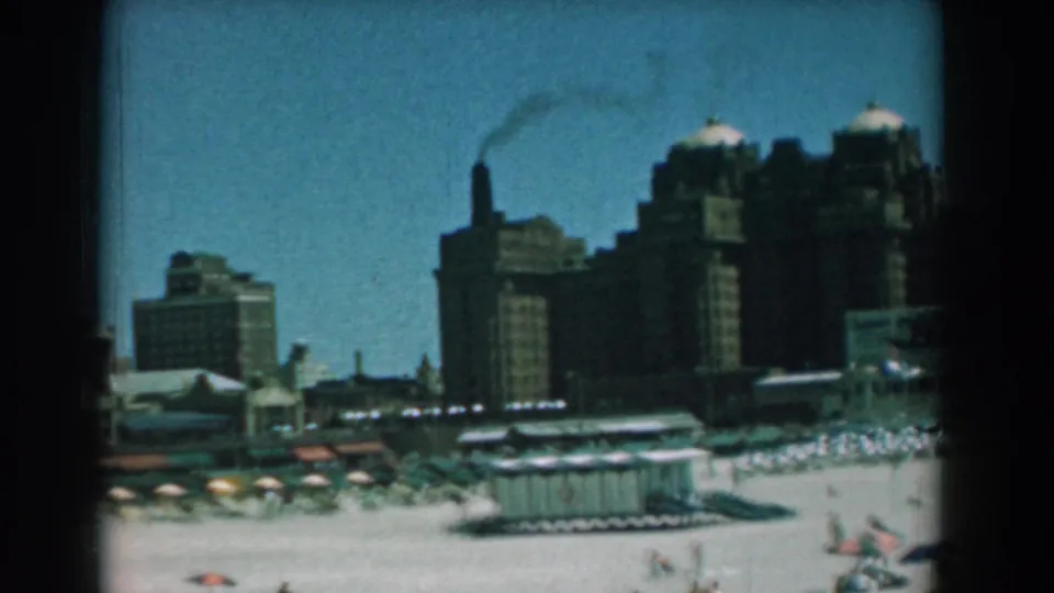 1958 Yorktown beach view with scattered bathers, colorful umbrellas, a low shore hut, and large buildings near a smoking stack.