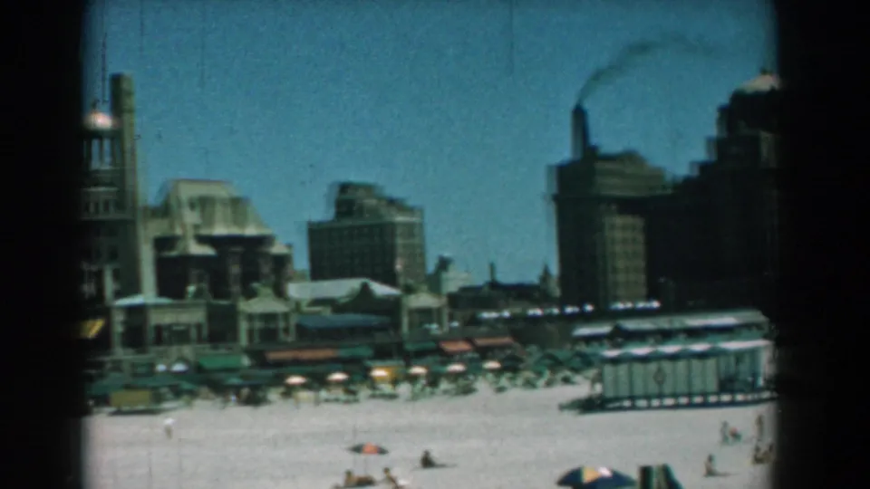 1958 Yorktown shoreline frame showing beach umbrellas, bathers, low huts, tall waterfront structures, and smoke rising at right.